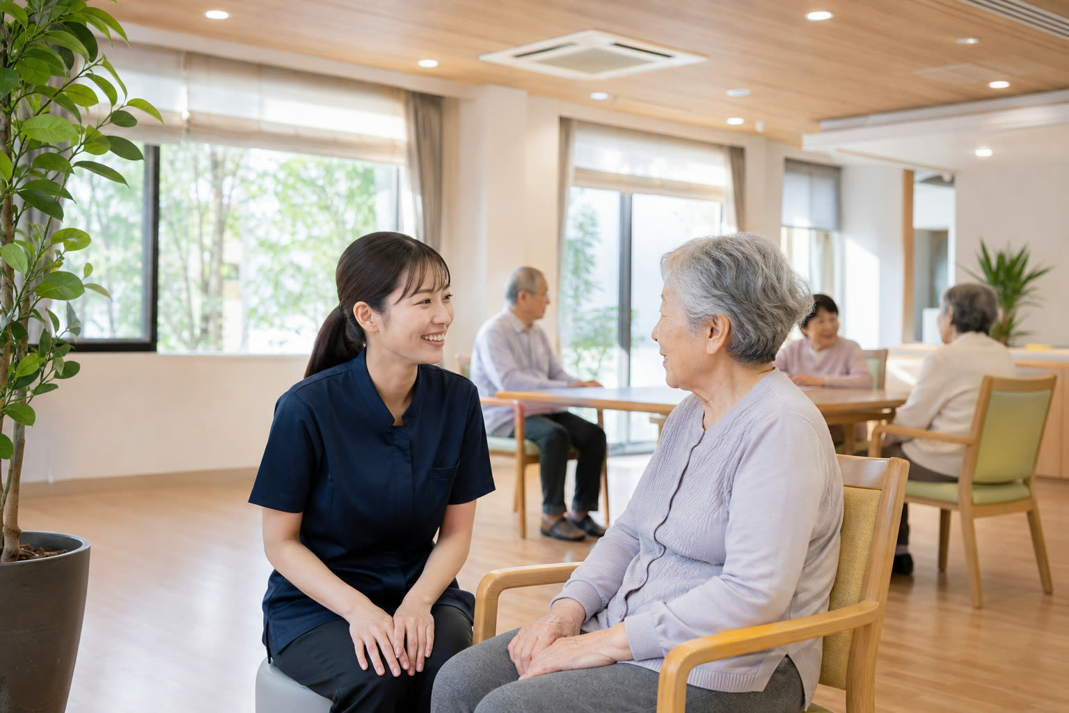 日中の共有フロアで入居者と話す紺色ユニフォームの介護スタッフ